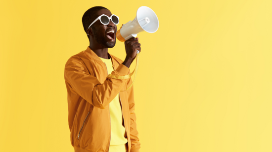 Ein Mann steht vor gelbem Hintergrund und ruft etwas in ein Grammophon, welches er in der Hand hält.